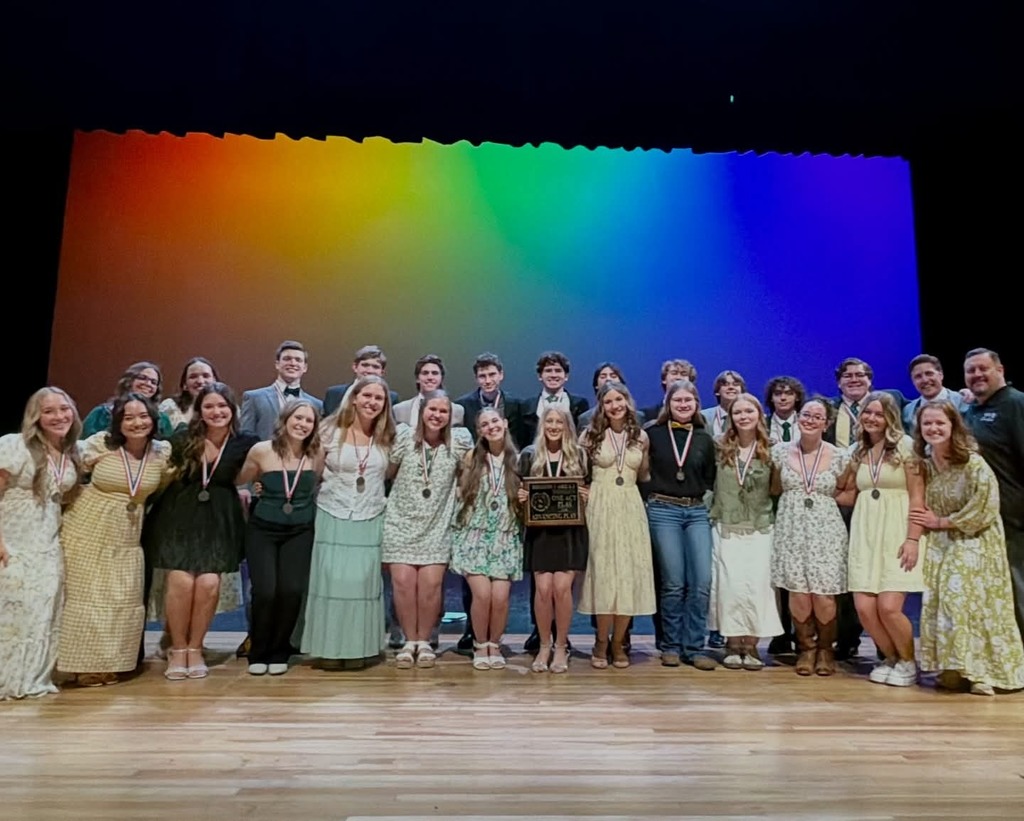 A large group of students and staff pose together on stage beneath a rainbow-colored theater backdrop, smiling after receiving medals and a plaque. The group is dressed in a mix of formal and semi-formal outfits, celebrating a team achievement at an academic or fine arts competition.