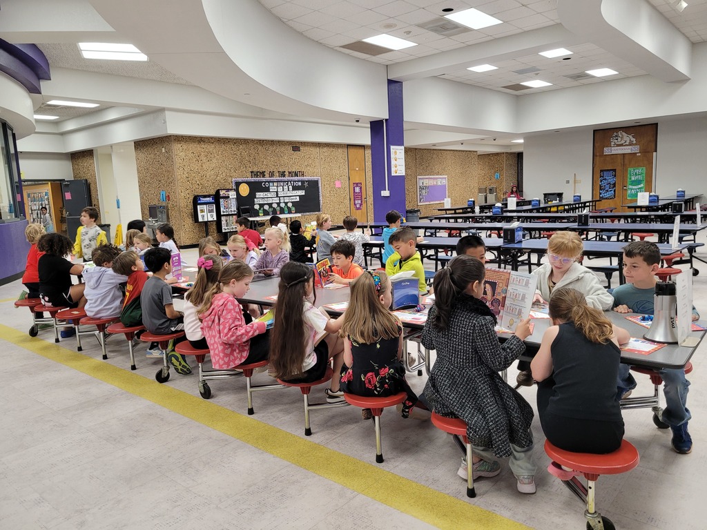 Students at Wylie West Elementary sit together in the cafeteria during a library celebration, reading books and visiting with classmates at lunch tables.