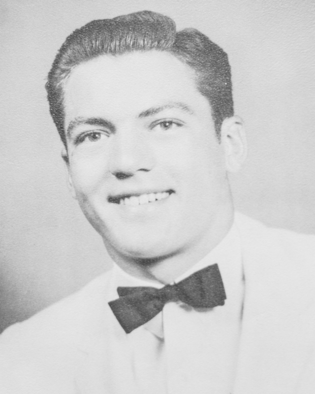 Black-and-white portrait of a young man wearing a light-colored suit jacket and bow tie, smiling at the camera with neatly styled hair in a vintage yearbook-style photo.