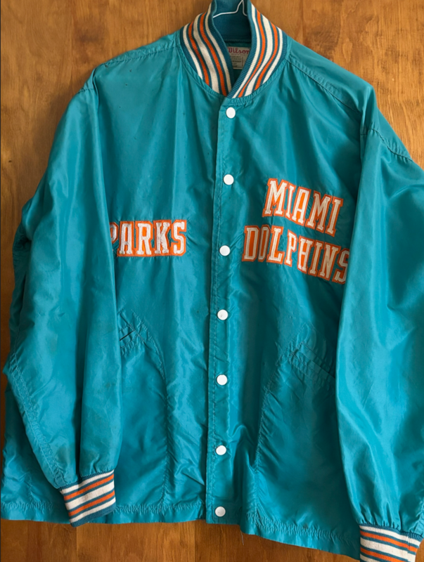 Teal Miami Dolphins jacket with orange and white lettering, featuring the name โParksโ on one side and โMiami Dolphinsโ across the front, hanging against a wooden background.