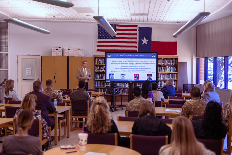 A presenter stands at the front of a library classroom speaking to a group of community members and military personnel seated at tables, with a screen displaying AFJROTC curriculum and U.S. and Texas flags hanging behind him.