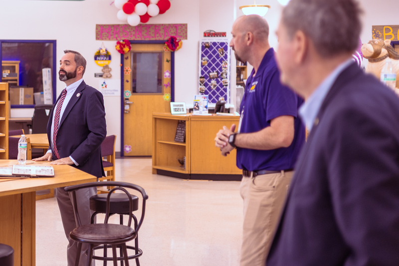 A speaker addresses a small group inside an elementary classroom, with school staff and visitors standing nearby as colorful decorations and a teacher’s door labeled “Mrs. Meek” are visible in the background.