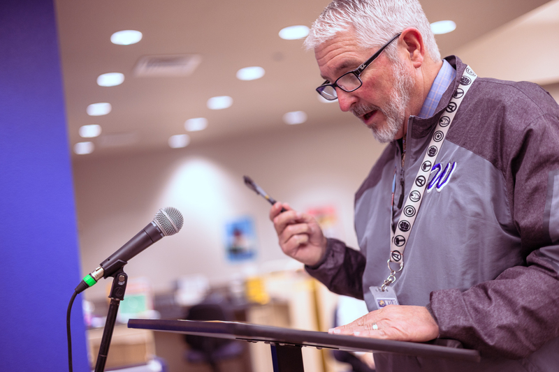 A man wearing a Wylie jacket and lanyard speaks at a podium during a meeting, holding a pen and leaning toward a microphone with a focused expression.