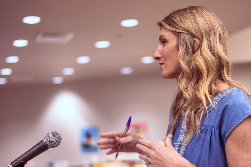 A woman stands at a microphone during a meeting, holding a pen and gesturing as she speaks in a school setting.