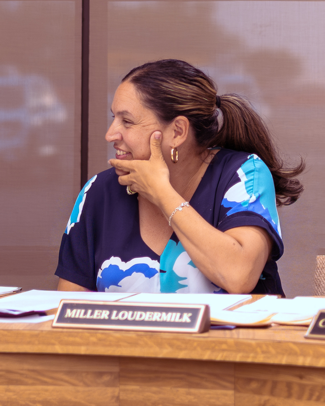 A Wylie ISD board member smiles while seated at the meeting table, listening with her hand resting near her face, nameplate visible in front of her.