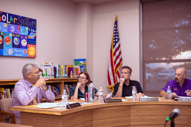 Members of the Wylie ISD Board of Trustees sit at a table during a meeting, listening and discussing items, with nameplates, water bottles, and an American flag visible in a school library setting.