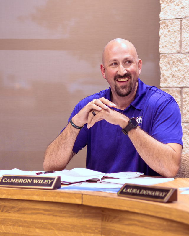 A Wylie ISD board member in a purple polo smiles during a meeting, seated at the table with papers and a nameplate in front of him.