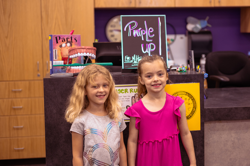Two kindergarten girls smile while standing in the Wylie East Elementary library. Behind them is the checkout desk with a colorful “Purple Up for Military Kids” sign, books, and library decorations.