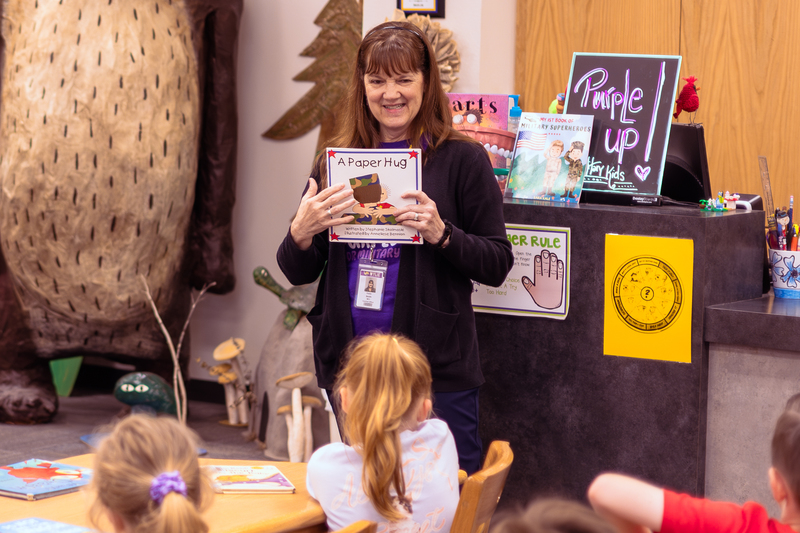 Mrs. Yvette Smith stands in the Wylie East Elementary library reading to kindergarten students while holding the book A Paper Hug. Behind her are books, a “Purple Up for Military Kids” sign, and library displays as students listen from tables.