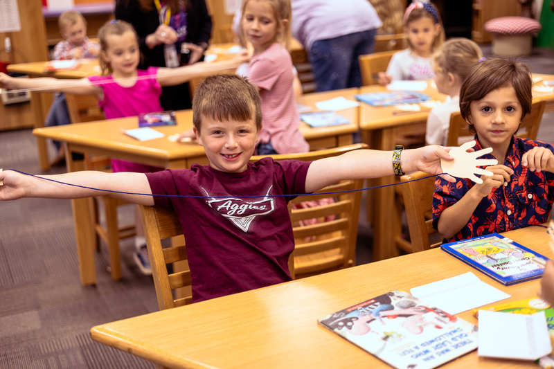 Kindergarten students participate in a classroom activity at tables in the Wylie East Elementary library while staff members assist around the room.