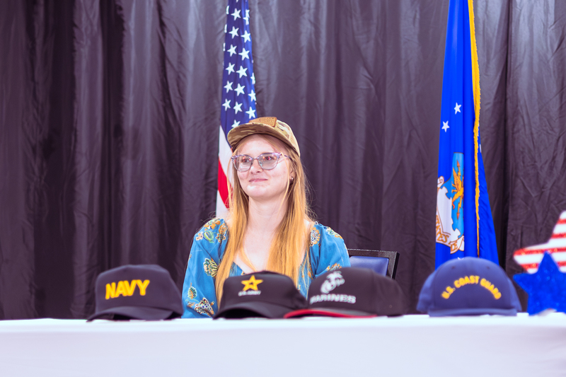 Student seated at a table looking upward during the signing ceremony with military hats in front.