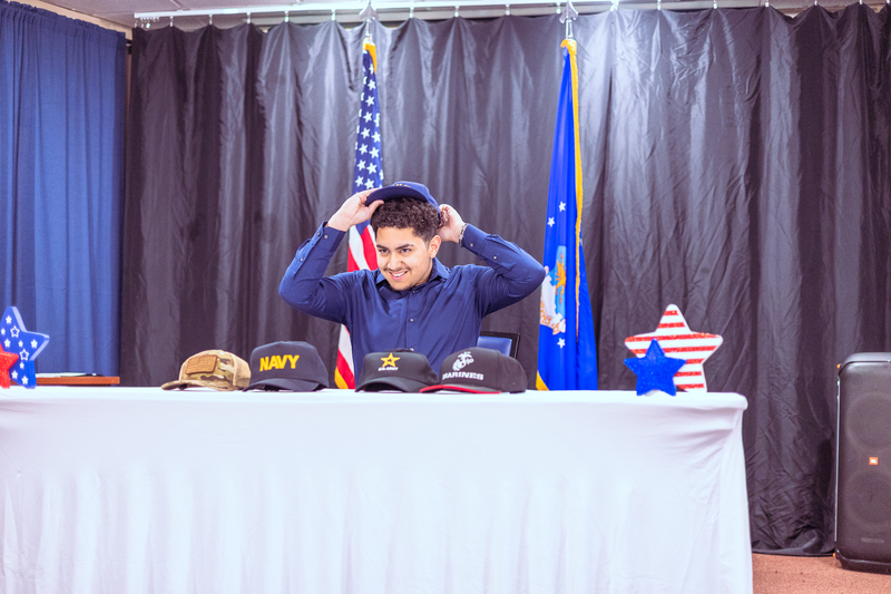 Student placing or adjusting a military branch hat on their head during the signing ceremony.
