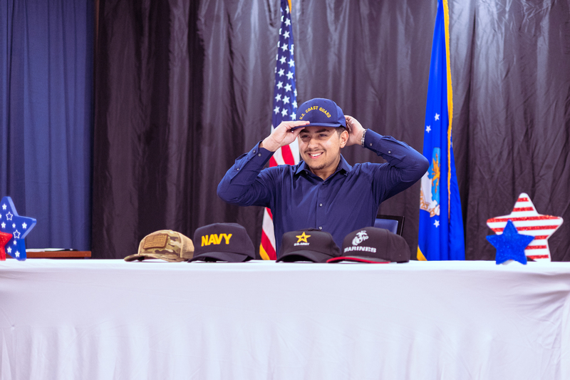 Student signing paperwork at a table during Military Signing Day with military flags in the background.