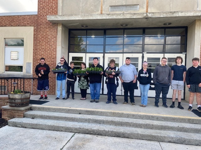10 students and 1 staff standing in front of the high school doors holding plants