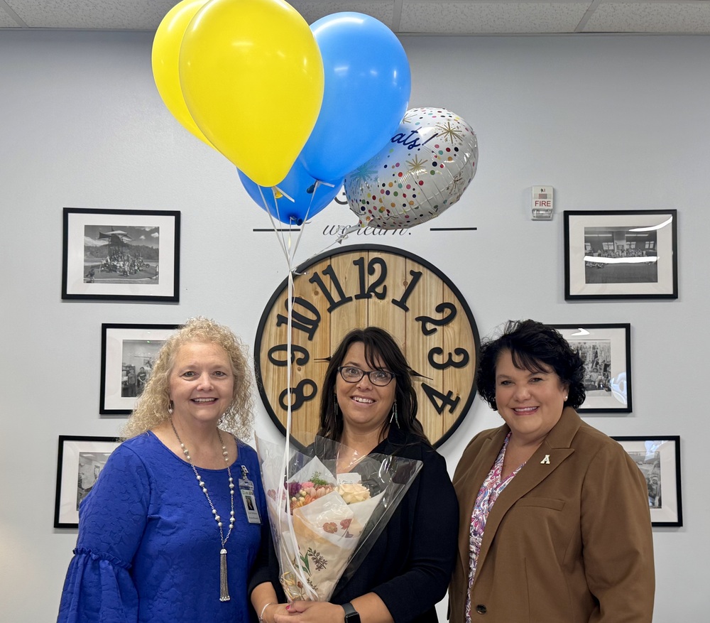 three women smiling. the woman in the middle is holding flowers and balloons