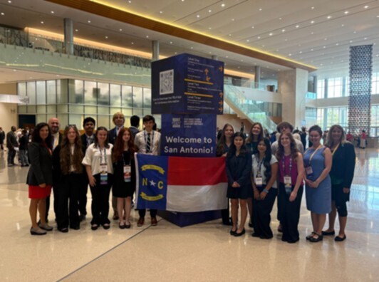 students and teachers in business attire standing on either side of NC flag with a sign that says "Welcome to San Antonio"