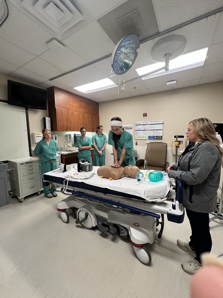girl in scrubs performing cpr on a dummy while a teacher and 3 students look on