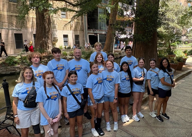group of students and teachers in light blue shirts on sidewalk in front of building