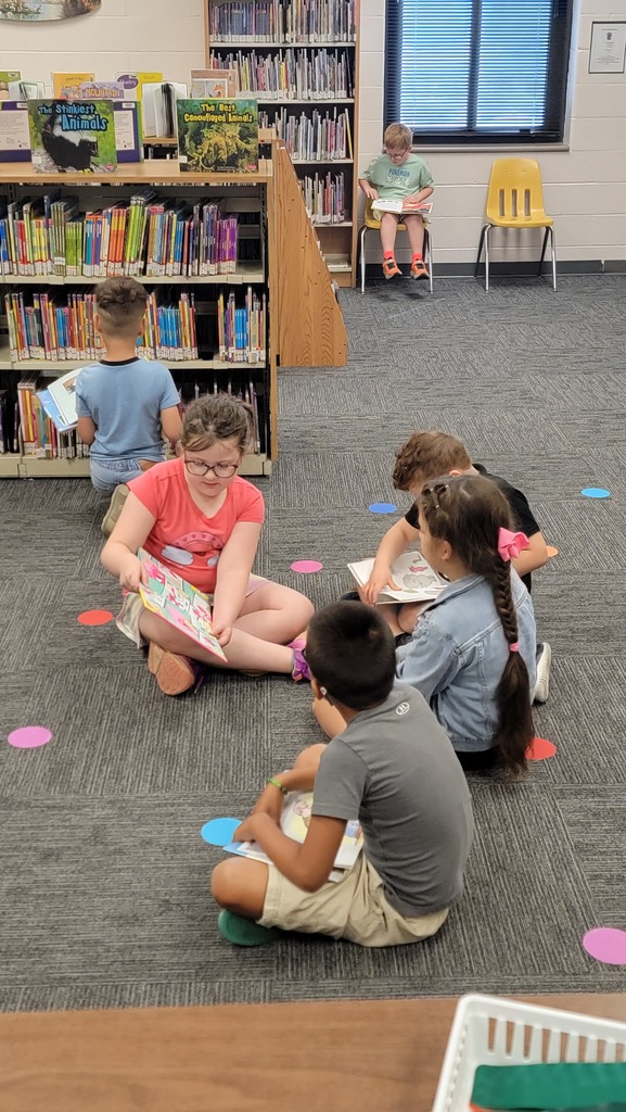 The picture shows children in the Lincoln School Library. Three children are sitting on the floor listening to a story being read to them by a classmate. In the background there are bookshelves and students looking quietly at books.