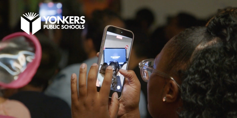 A parent records a student presentation on a smartphone during the Yonkers Middle High School Summer Enrichment Academy showcase.