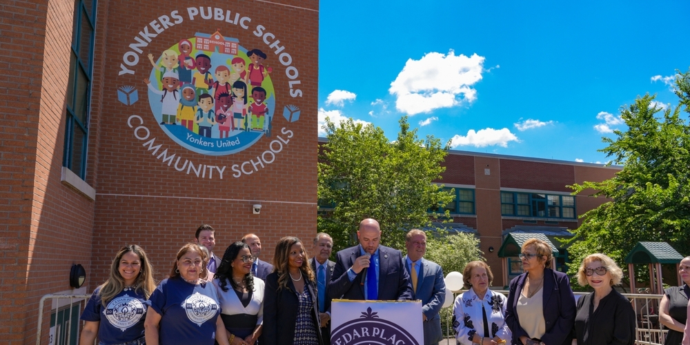 A group of school and community leaders stand outside Cedar Place Community School during a dedication ceremony. At center, Superintendent Aníbal Soler, Jr. speaks at a podium displaying the Cedar Place Community School emblem. Staff members, elected officials, and supporters stand alongside him beneath a bright blue sky. Behind the group, a large Yonkers Public Schools Community Schools mural featuring diverse students is prominently displayed on the brick school building. The scene reflects a celebratory moment marking the school's transition to Cedar Place Community School.