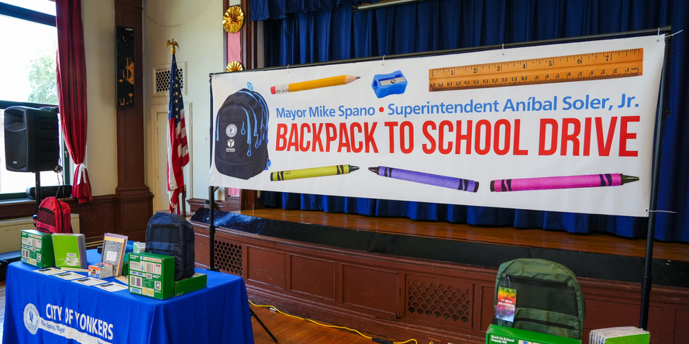 Promotional display for the City of Yonkers Backpack to School Drive inside an auditorium. A large banner featuring a backpack and school supplies spans the stage behind a table displaying donated backpacks and school supplies for students.