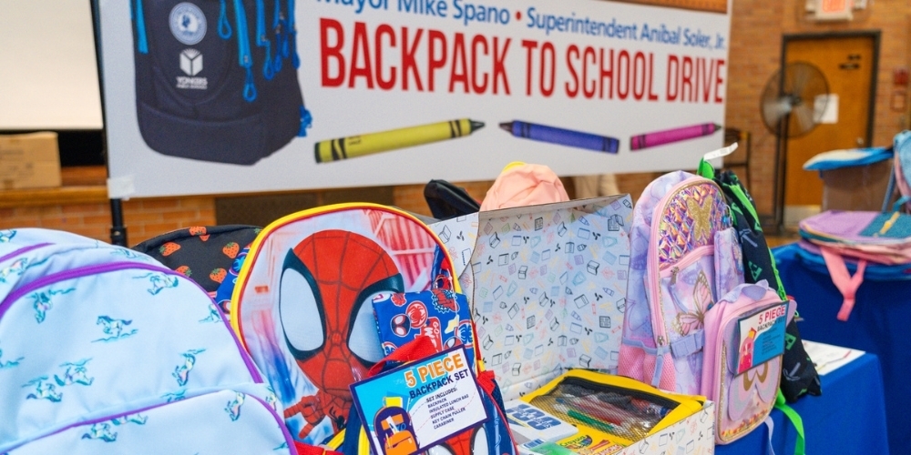 Beneath a large sign reading Mayor Mike Spano and Superintendent Aníbal Soler Jr Backpact to School Drive. a pile of colorful backpacks wait for young students to claim them. Prominent among the backpacks is one illustrated with Spiderman.