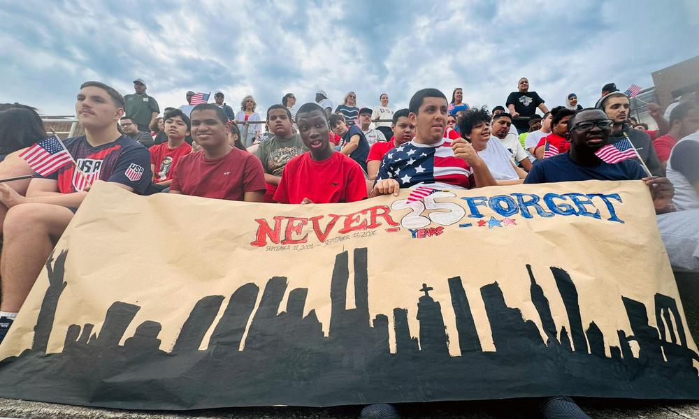 Roosevelt High School A-Team students hold American flags and a hand-painted “Never Forget — 25 Years” banner featuring the New York City skyline.