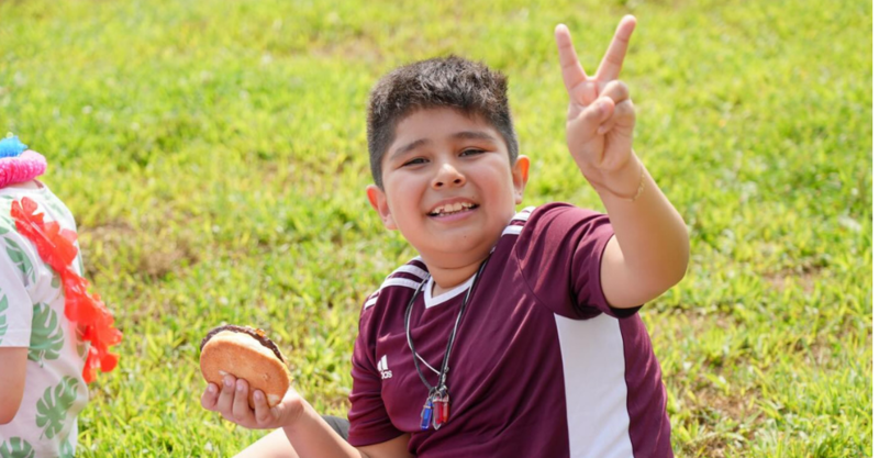Young boy flashing a V for Vidtory sign before he takes a bite out of his freshly grilled  burger