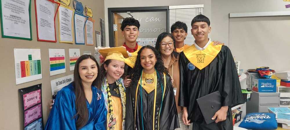 A group of smiling students and a teacher pose together inside a classroom during a graduation celebration. Several students wear black and gold graduation gowns and stoles, while others wear school spirit attire. One student holds a graduation cap. Classroom posters, schedules, and educational materials are displayed on the walls and tables in the background. The group stands closely together, celebrating their achievements and school pride.
