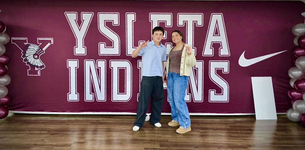 Two Ysleta High School students, a young man in a light blue shirt and a young woman in a beige cardigan, standing and smiling in front of a large maroon backdrop that reads “YSLETA INDIANS” with the YHS Indian mascot logo and a Nike swoosh. Both students are making the “Hook ’em Horns” hand gesture. The backdrop is flanked by maroon and silver balloon columns, and they are standing on a wooden floor.