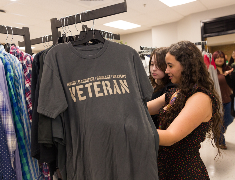 Two women look through clothes on racks. A gray shirt with the words Honor | Sacrifice | Courage | Bravery VETERAN is displayed in the foreground. Other shoppers and shirts are visible in the background.