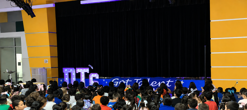 Students sit together in a school auditorium facing a stage decorated with illuminated “DT's” letters and a blue “Got Talent” banner. A large black stage curtain is centered between bright yellow walls with white trim. The audience of elementary students fills the foreground while the stage is prepared for a school talent show performance or presentation.