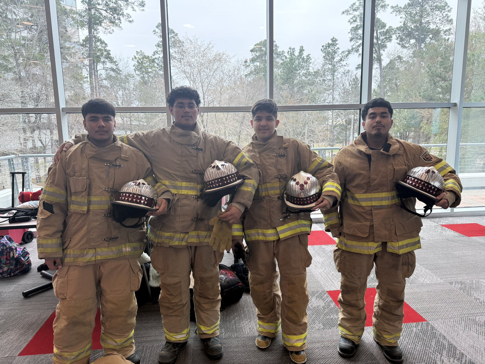 Group photo of four Ysleta High School students from the fire science or explorer program, dressed in full tan firefighter turnout gear with yellow reflective stripes, yellow trim, and black boots. They stand side-by-side with arms around each other's shoulders in a large, bright indoor atrium or conference hall featuring tall windows, pine trees visible outside, and a high ceiling. Each student holds a custom-decorated firefighter helmet: the helmets are silver with red and black designs, including patterns of triangles, arrows, and tribal-inspired motifs. The students appear proud and smiling, with luggage, backpacks, and gear scattered on the floor behind them, suggesting they are attending a competition, conference, or training event. The setting includes natural light from large glass walls overlooking a wooded area.