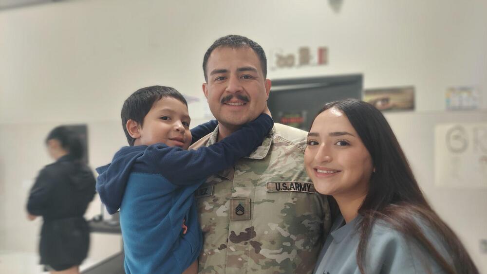 A smiling U.S. Army soldier in camouflage uniform stands between a young boy and a young woman for a family photo indoors. The boy, wearing a blue hoodie, hugs the soldier around the neck, while the woman with long dark hair leans close to the soldier. All three are smiling at the camera. The background is softly blurred and appears to be an indoor public or military facility.