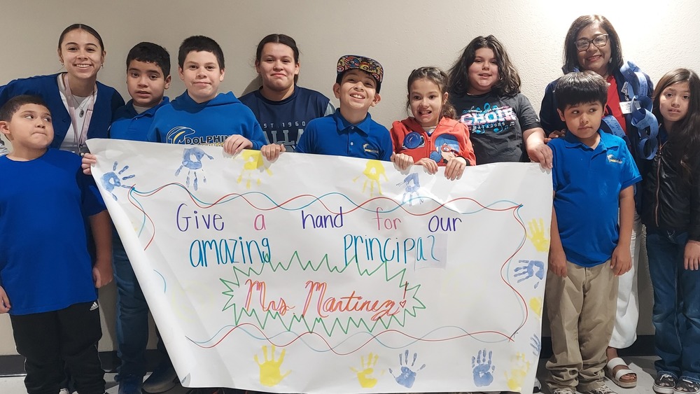 Students and staff from Dolphin Terrace proudly gathered together to celebrate Principal Appreciation Day with Mrs. Martinez. Surrounded by smiling students, the group held a colorful handmade banner reading, “Give a hand for our amazing principal,” decorated with bright handprints and cheerful designs. The photo captures the strong sense of community, appreciation, and school spirit that Mrs. Martinez inspires every day through her leadership, kindness, and dedication to students and families.