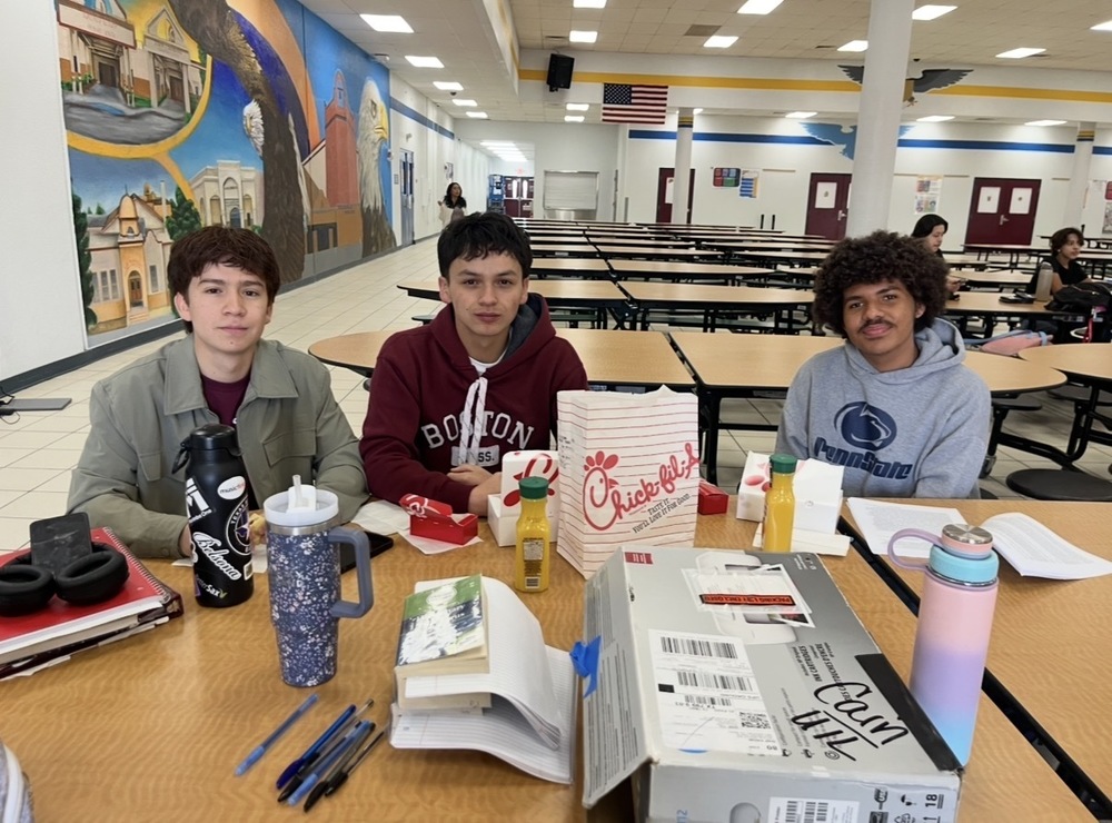 Three teenage boys sitting at a lunch table in a school cafeteria, smiling at the camera. The boy on the left wears a green jacket and has a black water bottle and earbuds; the middle boy wears a maroon "Boston" hoodie; the boy on the right wears a gray Penn State hoodie and has curly hair. On the table are Chick-fil-A food, drinks, notebooks, pens, and a large cardboard box. In the background, there's a colorful mural with eagles and buildings, empty cafeteria tables, and an American flag.