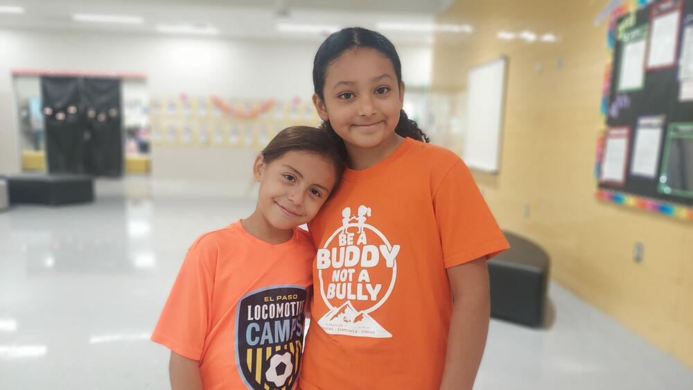 Two students stand together in a school hallway, smiling at the camera. Both are wearing orange T-shirts to promote kindness and anti-bullying awareness. The older student’s shirt reads “Be a Buddy, Not a Bully,” while the younger student’s shirt features an El Paso Locomotive FC Camps design. The students pose with their heads gently leaning together, conveying friendship and school unity. The background features classroom displays, bulletin boards, and school furniture.