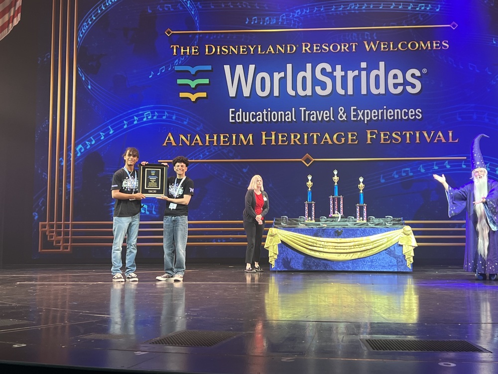 2 Choir students hold an award plaque together, standing on a stage. A large screen behind them reads The Disneyland Resort Welcones WorldStrides Educational Traves and Experiences Anaheim Heritage Festival. A trophy table with gold and blue decorations is in the background.