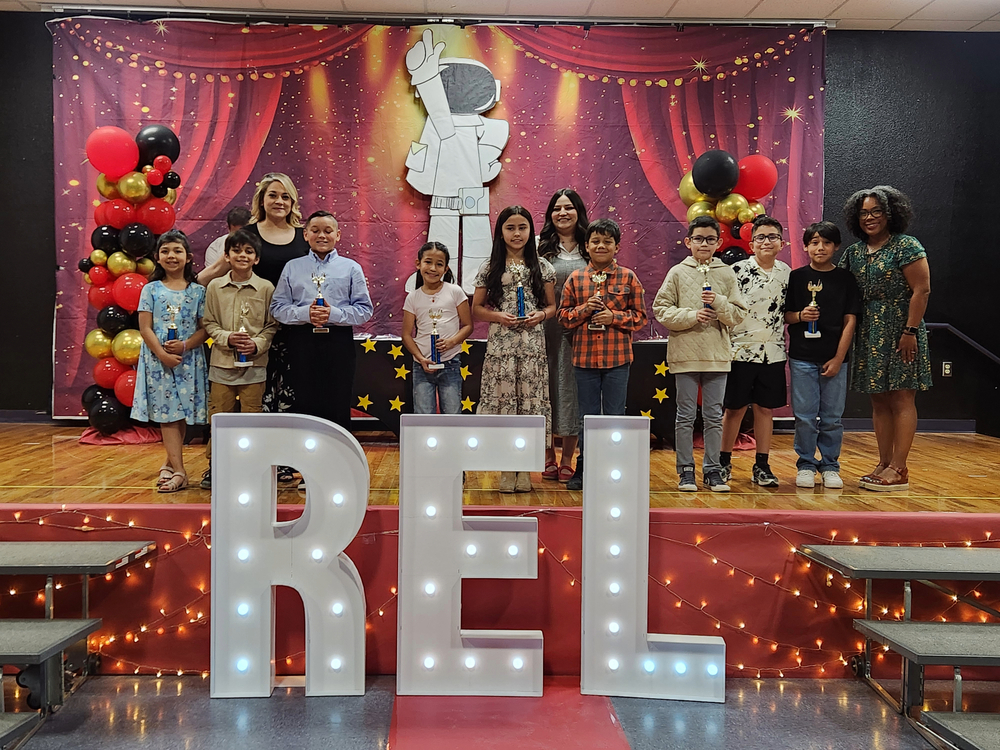 Several 3rd grade students holding their trophies and standing on stage with their teacherse after receiving their award. 