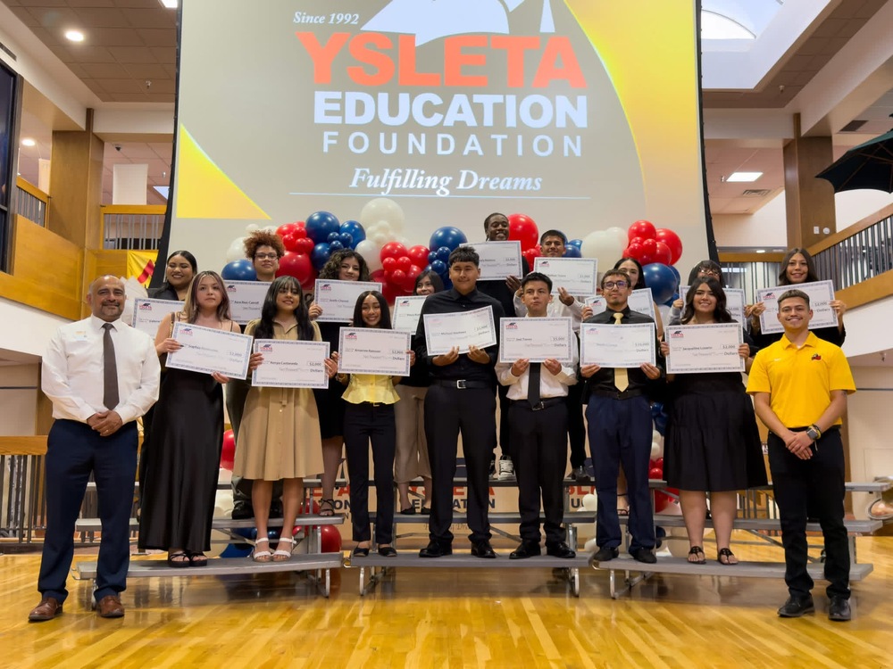 Here is a detailed alternative text description for the image, optimized for web accessibility: **Alt Text:** A group photo on an indoor stage at the Ysleta Education Foundation ceremony. Sixteen high school students stand on a multi-tiered riser, holding oversized scholarship checks. They are flanked by two men: a school or district official on the far left wearing a white shirt, tie, and dark pants, and another official on the far right wearing a yellow polo shirt and black pants. A massive projector screen behind the group reads "Since 1992, YSLETA EDUCATION FOUNDATION, Fulfilling Dreams" in large orange and white text, framed by red, white, and blue balloons. The students are dressed in semi-formal attire, and the overall atmosphere is celebratory.