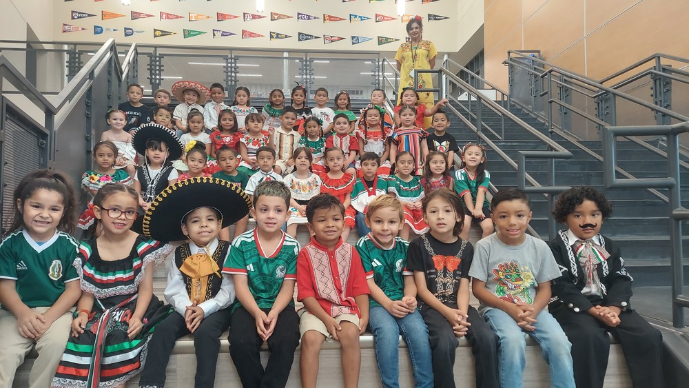 A large group of elementary-aged students pose together on a set of indoor school bleachers. The students are arranged in several rows and are smiling toward the camera. Many are wearing colorful traditional Mexican clothing, including embroidered dresses, decorative shirts, sombreros, serapes, and accessories in red, green, white, and other bright colors. Several students wear Mexico-themed shirts and outfits. University pennants and banners are displayed along the upper wall behind the group, creating a school-themed background.