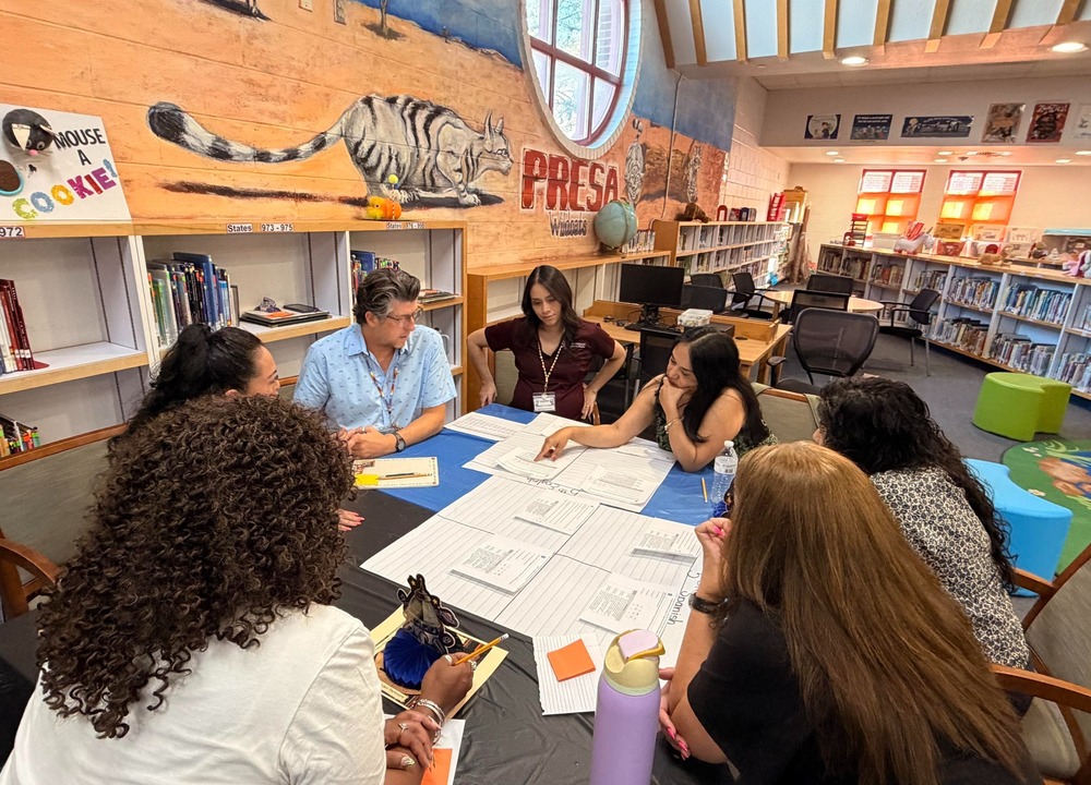 Presa teachers gathered around a library table working on collecting data the Presa Wildcat and Name are on the backwall under the the round window