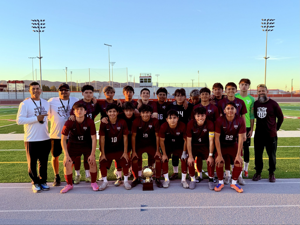 A large group photo of the Ysleta High School boys' soccer team (wearing maroon jerseys with “YHS” and numbers) posing proudly on a football/soccer field at sunset. The team is kneeling and standing in multiple rows with their 2026 4A Playoff championship trophy (a golden soccer ball on a pedestal) placed in the center foreground. Several coaches and staff members stand on the sides, including one in a white long-sleeve shirt pointing forward, another in a black cap, and one on the right in a maroon shirt with a beard. The players show team spirit with arms around each other, smiling at the camera. Stadium lights, bleachers, a scoreboard, and distant mountains are visible in the golden-hour background.