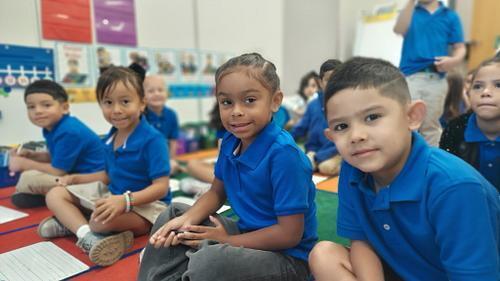 Male and Female students with blue shirts are sitting on a carpet in the classroom working on writing.