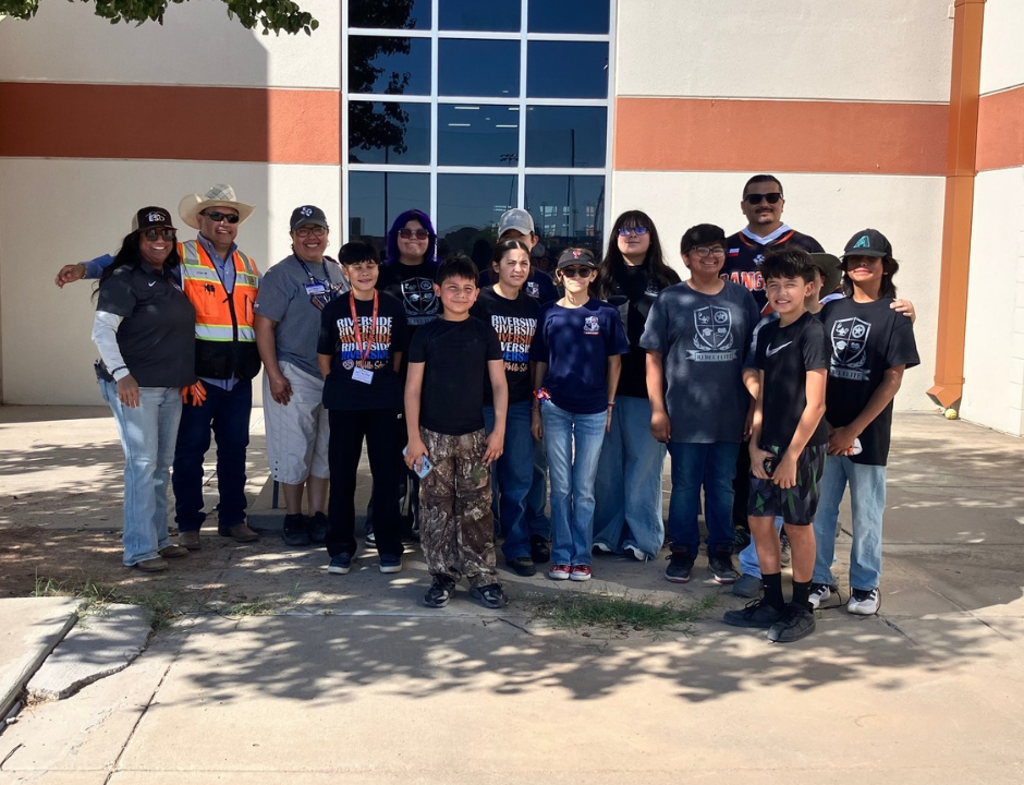A group of adults and students pose for a group photo outside a modern school or community building on a sunny day.