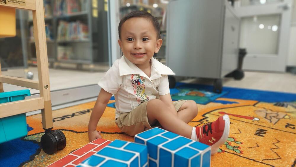 A young child sits on a colorful classroom rug surrounded by blue and red building blocks. The child is smiling toward the camera and wearing a white collared shirt with a colorful floral design, khaki shorts, white socks, and red sneakers. The classroom background includes bookshelves, storage furniture, educational materials, and a wooden activity shelf. The image captures the child engaged in a bright, playful classroom environment.