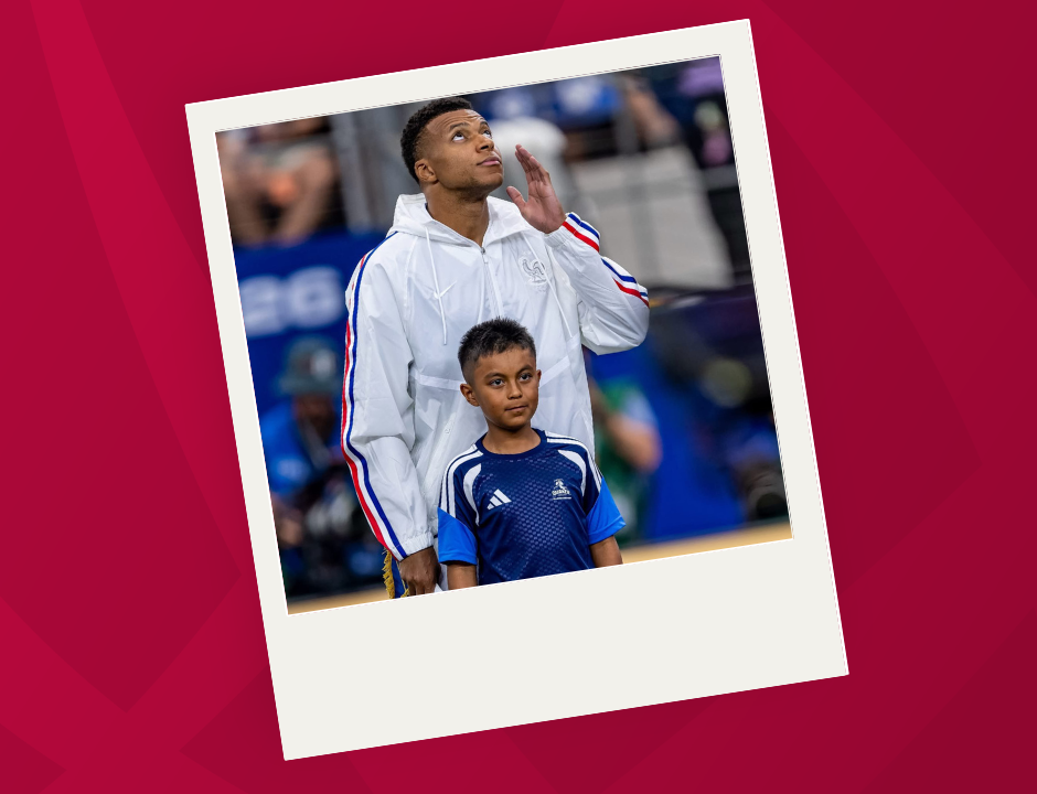 A male soccer player in a white jacket stands behind a young boy in a blue uniform on a field, with the player looking up and touching his face, during a pre-game ceremony.