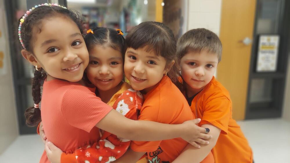 Four young children wearing orange shirts are standing close together in a group hug, smiling at the camera. One child wears a coral-colored shirt and a colorful beaded headband, while another has orange hair clips. The children are embracing one another in a warm display of friendship and unity in a school hallway.