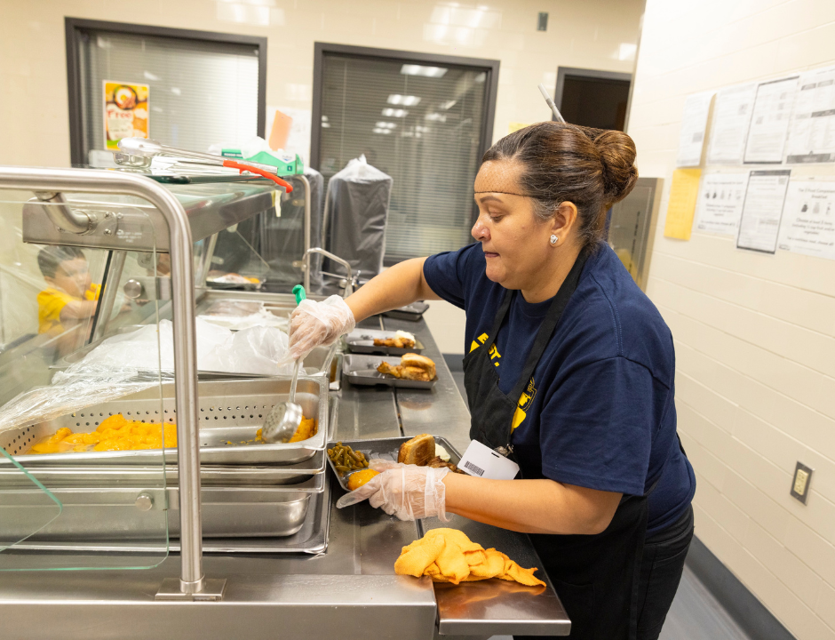 A cafeteria worker wearing gloves serves food, including fried items and vegetables, onto trays in a school kitchen.